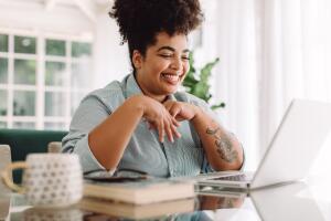 Mulher negra sorridente trabalhando em home office, sentada à mesa com um laptop, livros e uma caneca ao lado. O ambiente é iluminado e aconchegante, transmitindo conforto e produtividade no trabalho remoto.