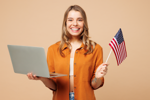 mulher branca sorridente, um notebook em uma mão e uma bandeira dos estados unidos na outra.