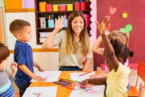 Imagem de uma sala de aula, a professora está com a mão levantada e um dos alunos também, todos com expressões felizes