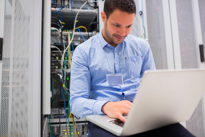 Homem feliz, sentado, com um notebook no colo. No fundo da imagem um servidor empresarial.