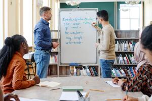 Uma aula de inglês em uma biblioteca, onde um professor e um aluno escrevem no quadro a conjugação de verbos regulares e irregulares em inglês. Outros alunos participam atentamente, anotando em seus cadernos.