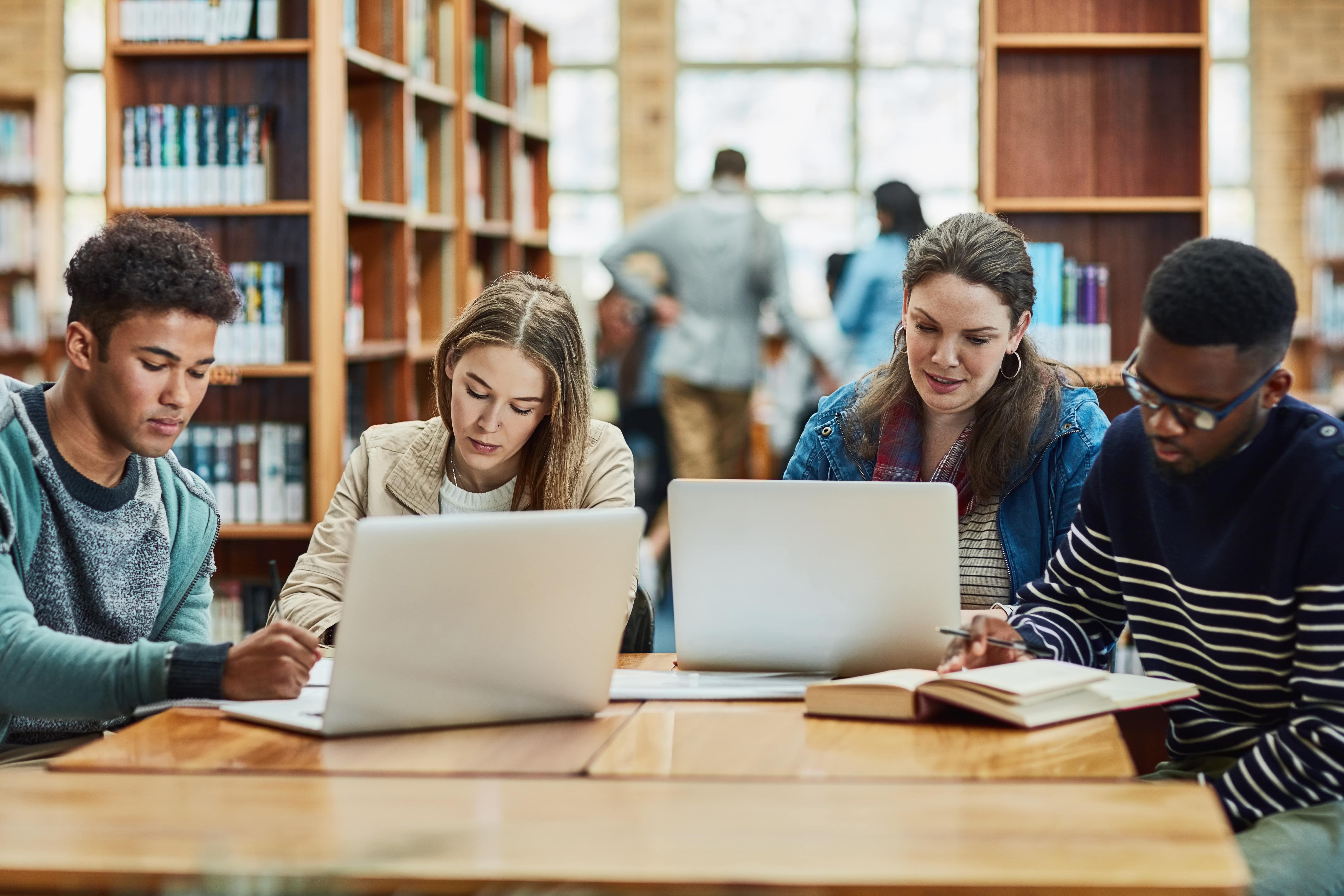 A imagem mostra quatro estudantes sentados em uma mesa de biblioteca, focados em atividades de estudo. Dois deles estão utilizando laptops, enquanto outros dois estão escrevendo em cadernos.