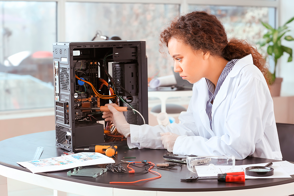 Mulher de jaleco branco sentada em uma mesa e muito concentrada em manipular uma cpu de computador.