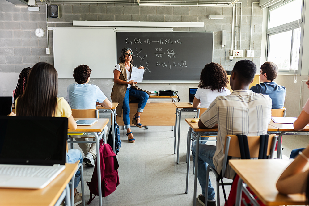 Uma foto de sala de aula, a professora ao fundo encostada em uma mesa, sorrindo.