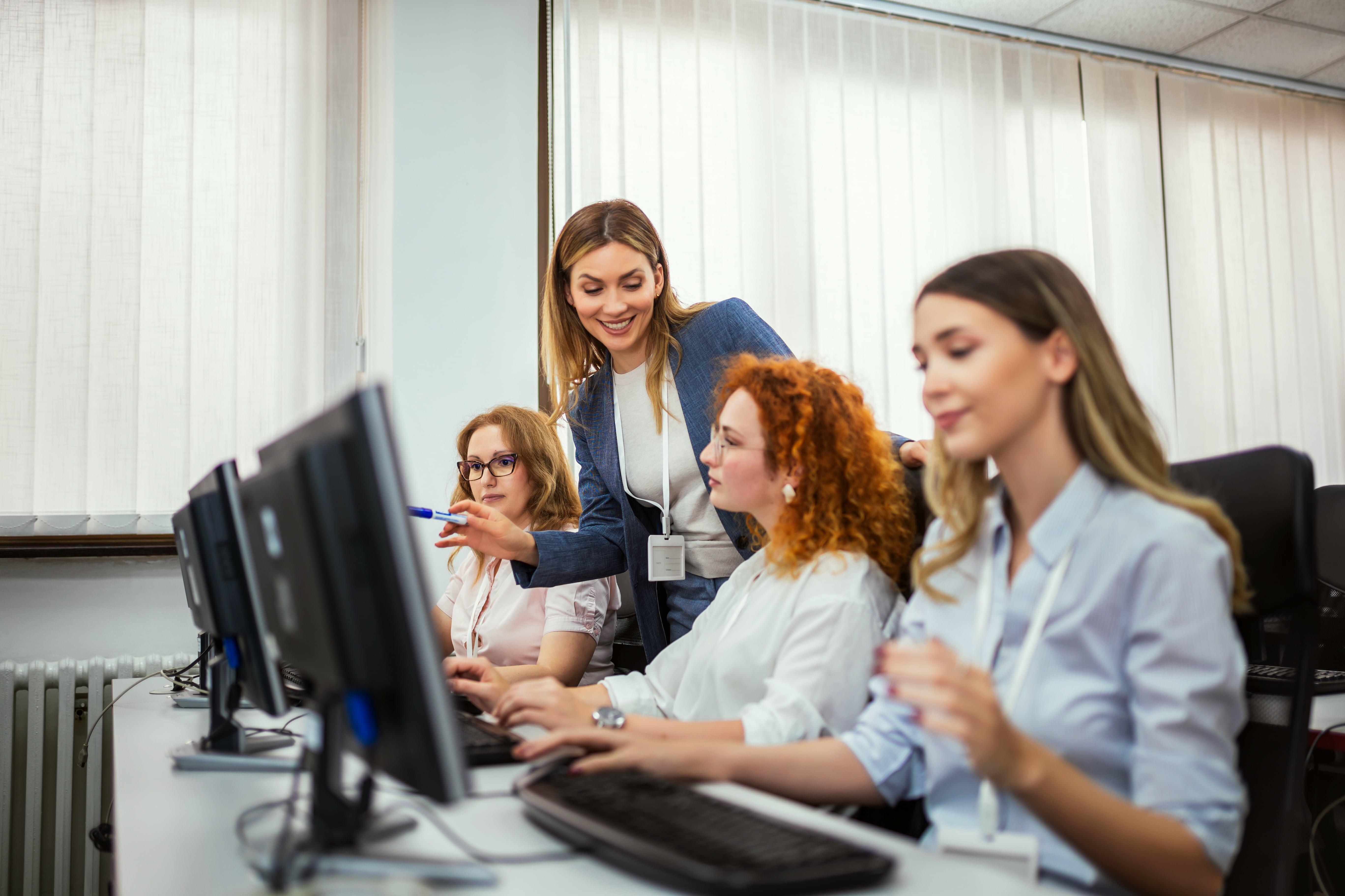 Aula prática de curso de informática com alunos aprendendo em um laboratório equipado. A instrutora orienta os participantes no uso do computador, destacando a importância da capacitação digital para o mercado de trabalho.