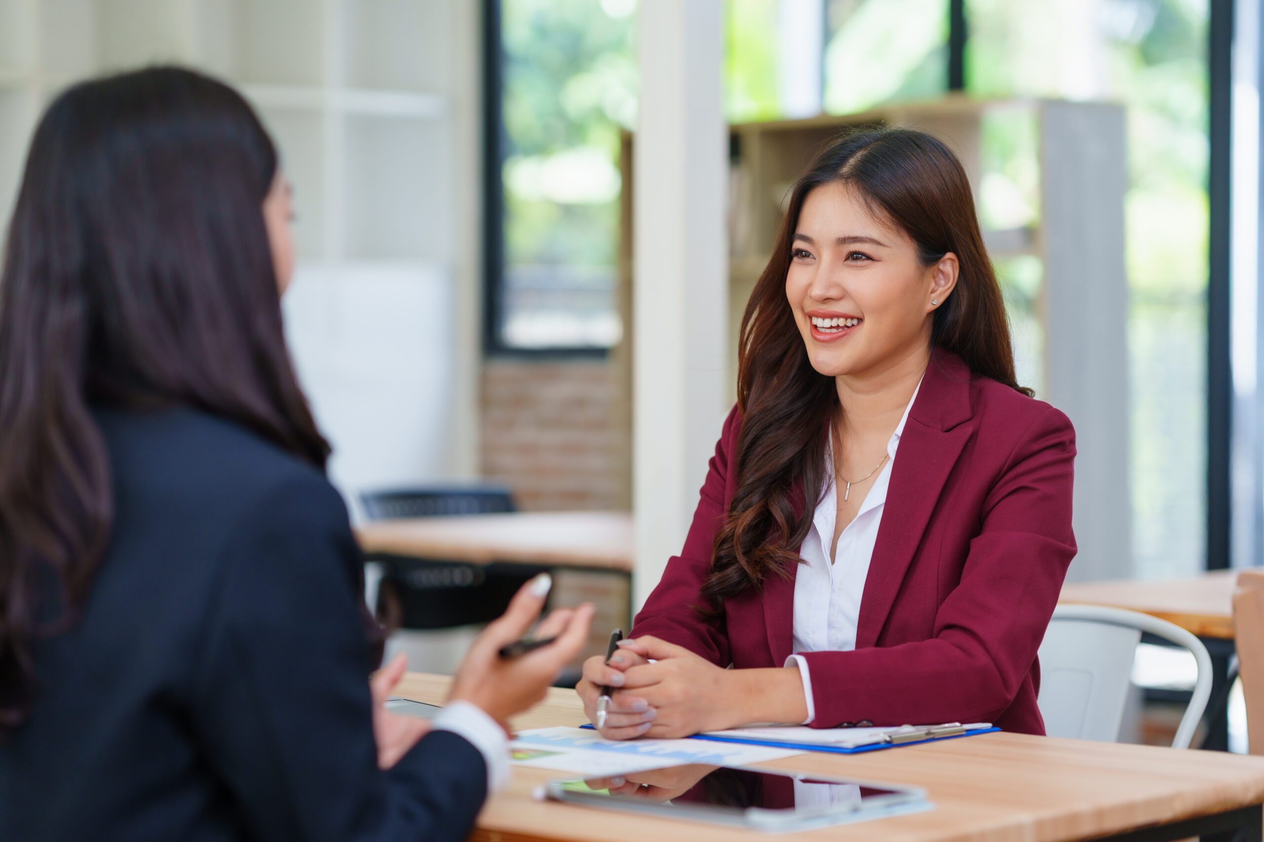 Duas mulheres em uma entrevista de emprego, sorrindo e discutindo em um ambiente de escritório moderno com janelas grandes.
