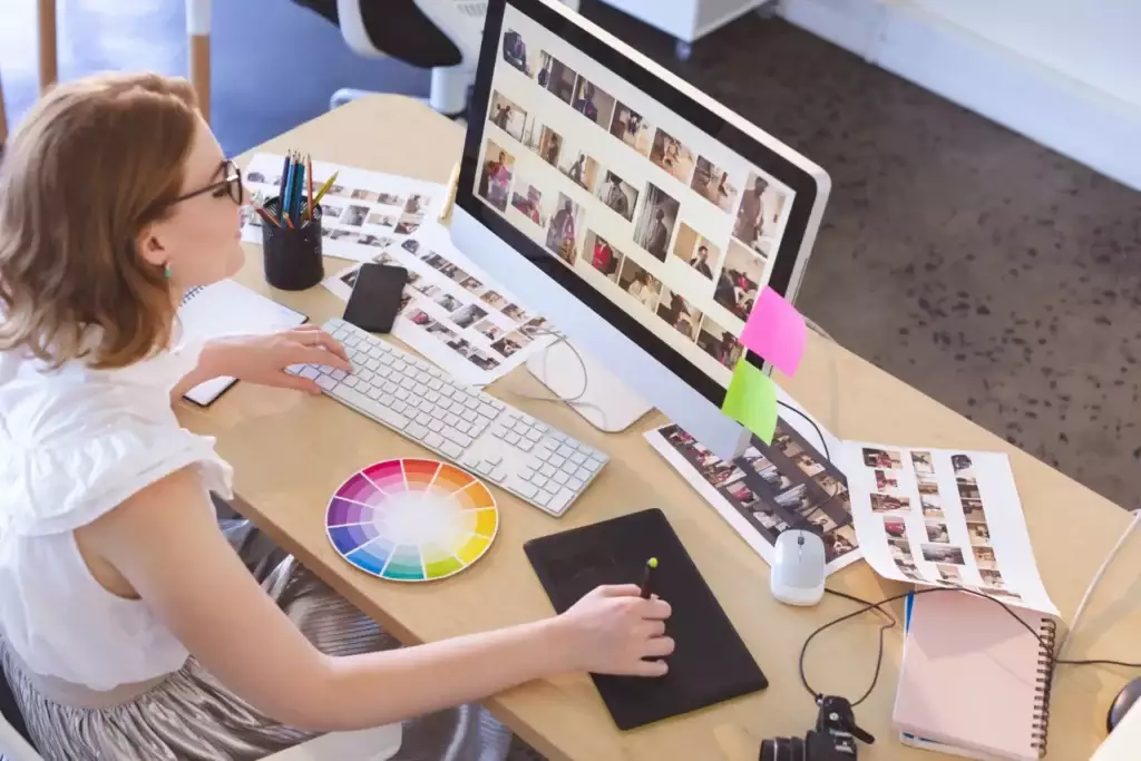 Designer gráfico mulher editando fotos em seu computador, com várias fotos impressas e materiais de trabalho na mesa, ilustrando um processo de edição de fotos.