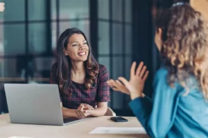 Mulher sorridente em entrevista de emprego, sentada em uma mesa com laptop, conversando com outra pessoa, ambiente de escritório moderno