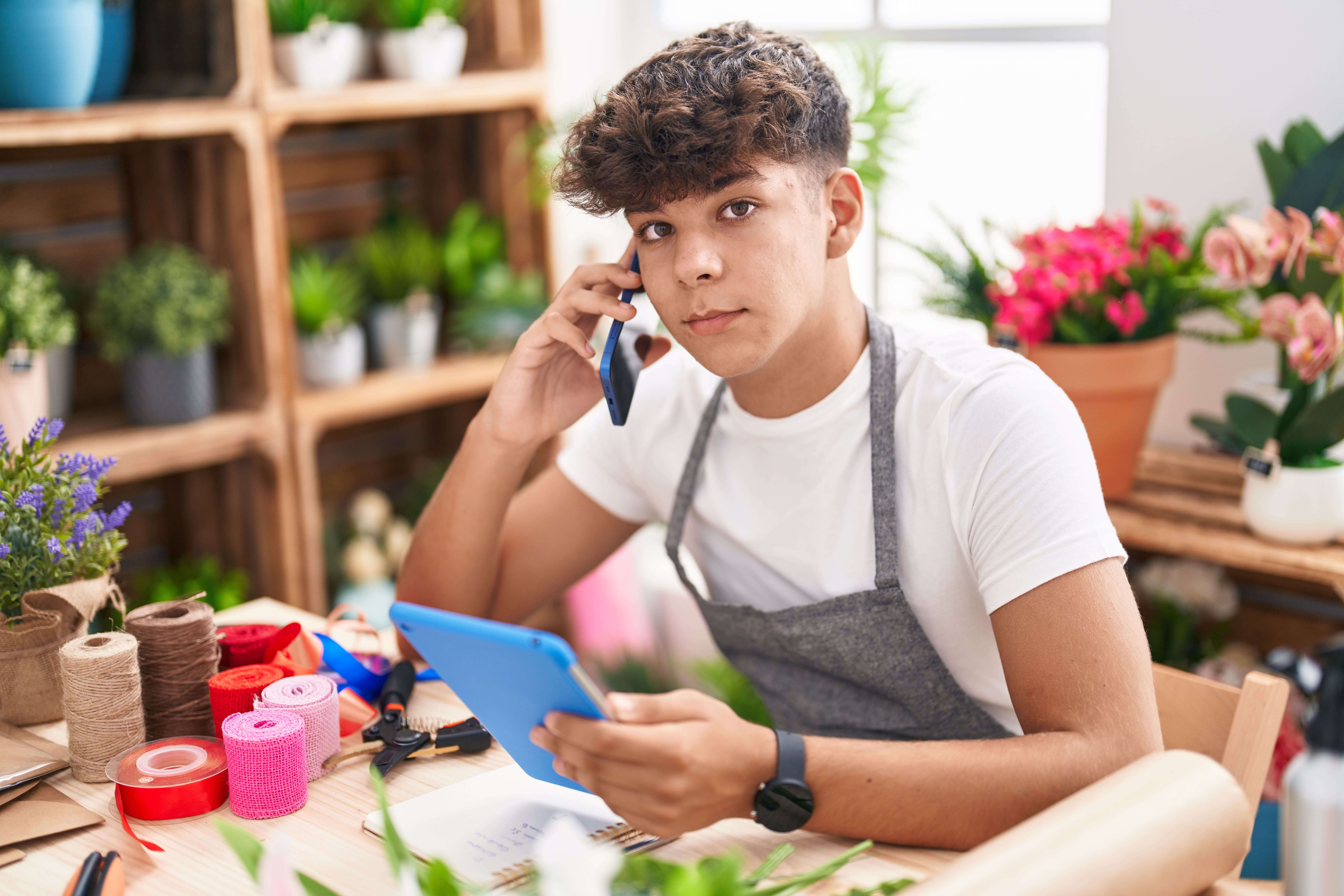 Jovem florista conversando ao telefone enquanto trabalha em sua loja de flores, rodeado de diferentes tipos de plantas e materiais agrícolas.