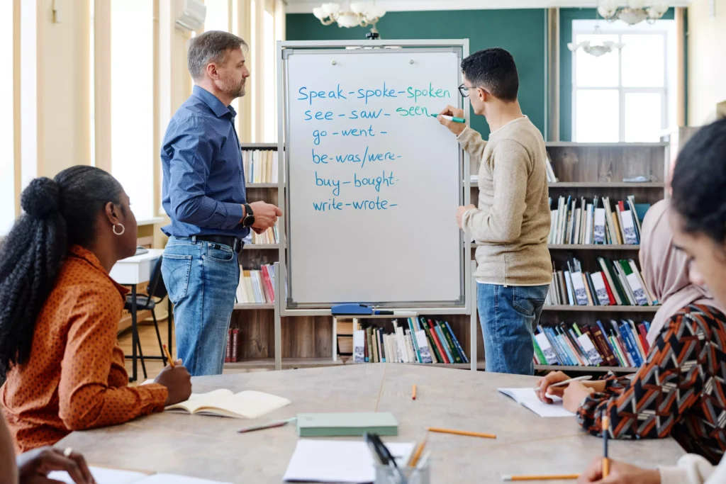 Aula de inglês com professor e estudantes praticando vocabulário de tempos verbais, como 'speak', 'saw', 'go', 'be', 'buy', e 'write', em uma sala de aula bem iluminada.