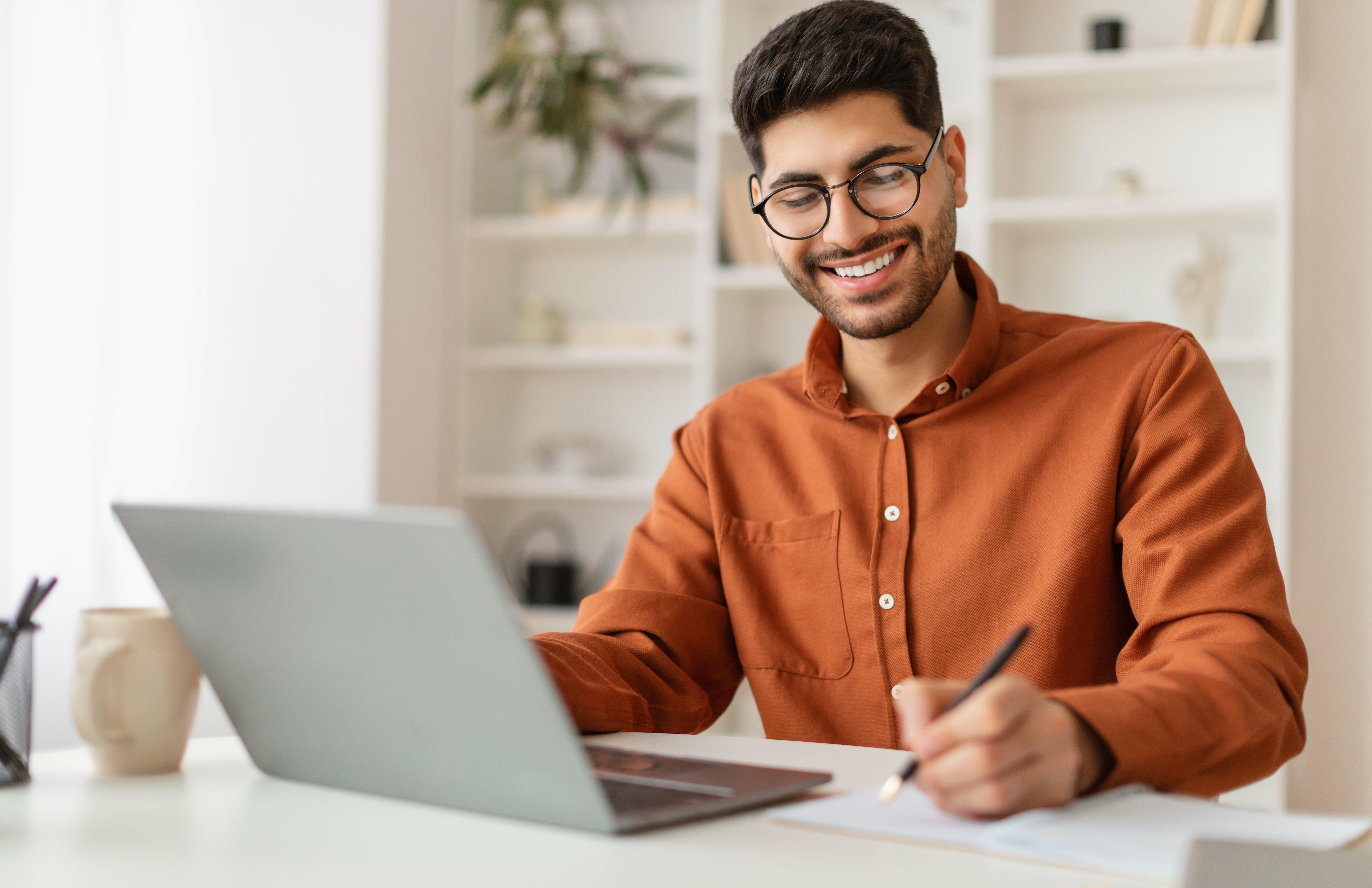 Jovem sorridente estudando em casa no computador e fazendo anota&ccedil;&otilde;es, representando os 4 passos para se conhecer melhor no dia a dia e a import&acirc;ncia do autoconhecimento na vida profissional