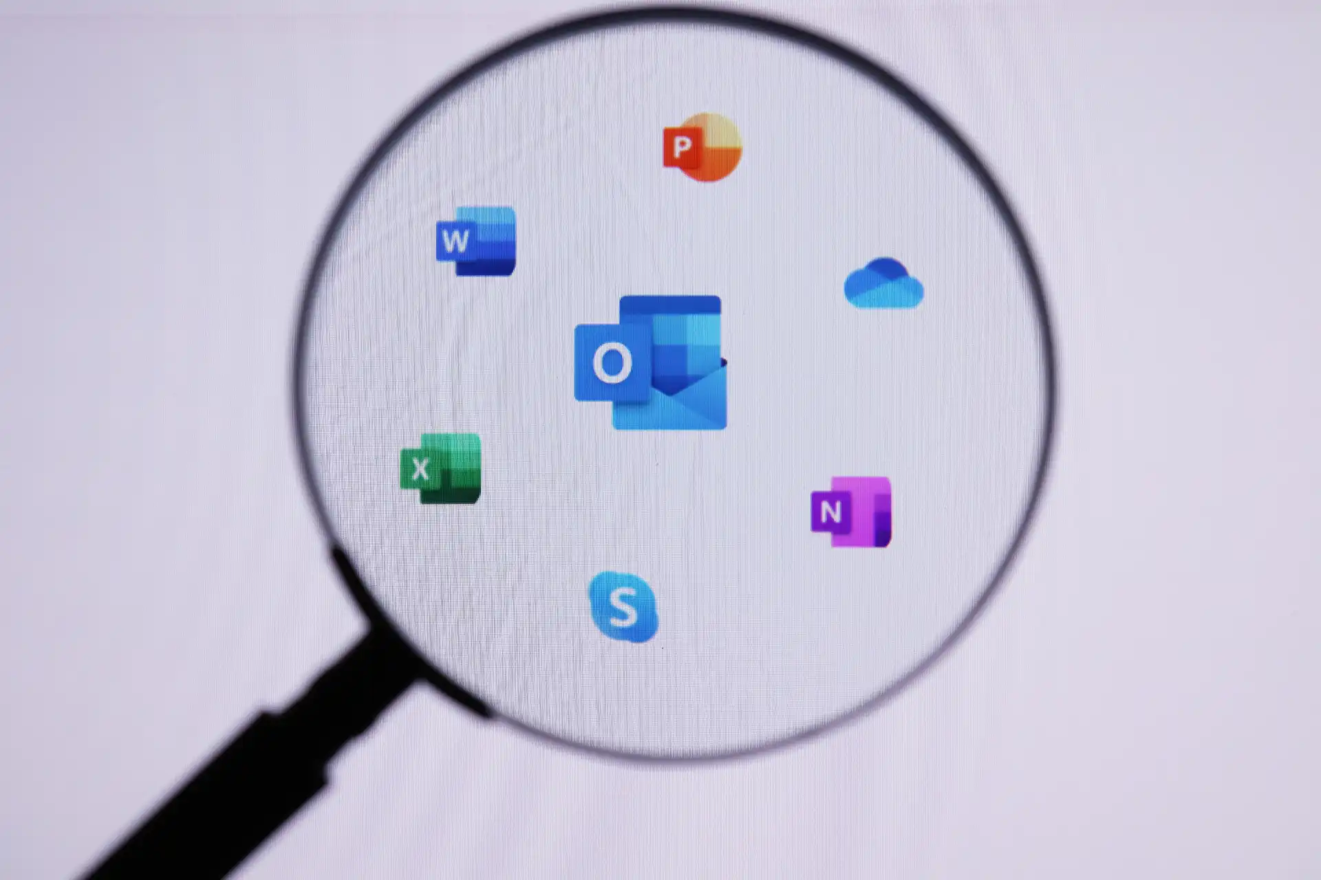 Imagem de uma lupa ampliando os ícones do pacote Microsoft Office, incluindo Word, Excel, PowerPoint, Outlook, OneNote, Teams e OneDrive, simbolizando programas de produtividade e colaboração digital.