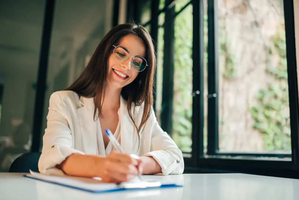 Mulher profissional em ambiente de escritório, sorrindo enquanto escreve em um documento com caneta azul, usando óculos, transmitindo foco e postura objetiva-para-currículo.