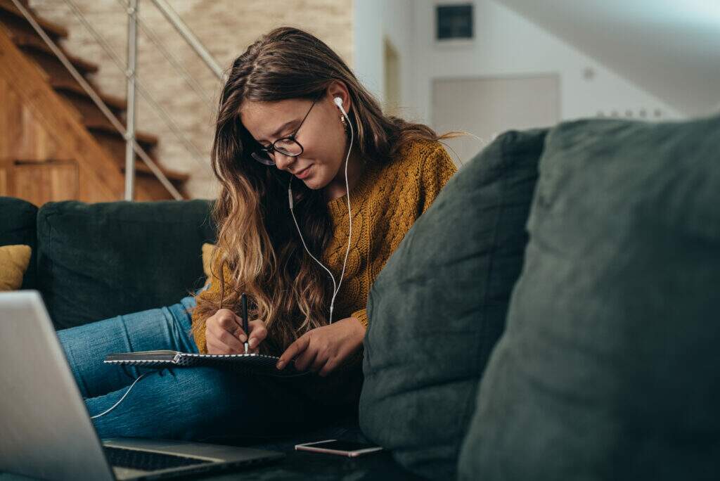 Jovem estudando de forma autodidata em casa, com caderno, computador e materiais de estudo organizados sobre o sofá.