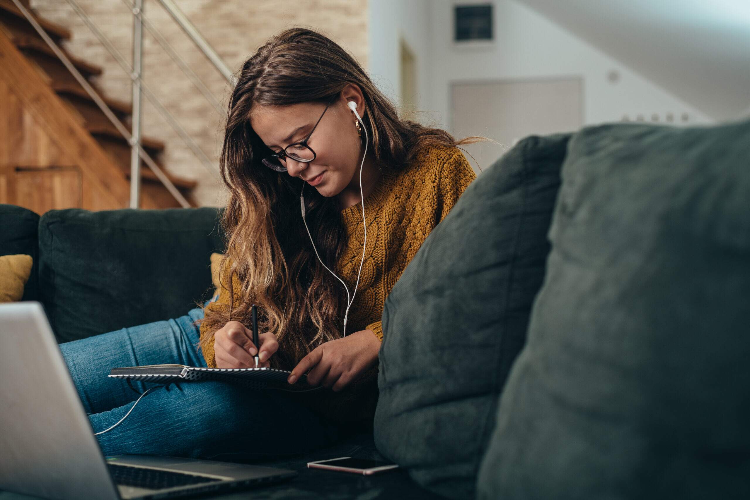 Jovem estudando de forma autodidata em casa, com caderno, computador e materiais de estudo organizados sobre o sofá.