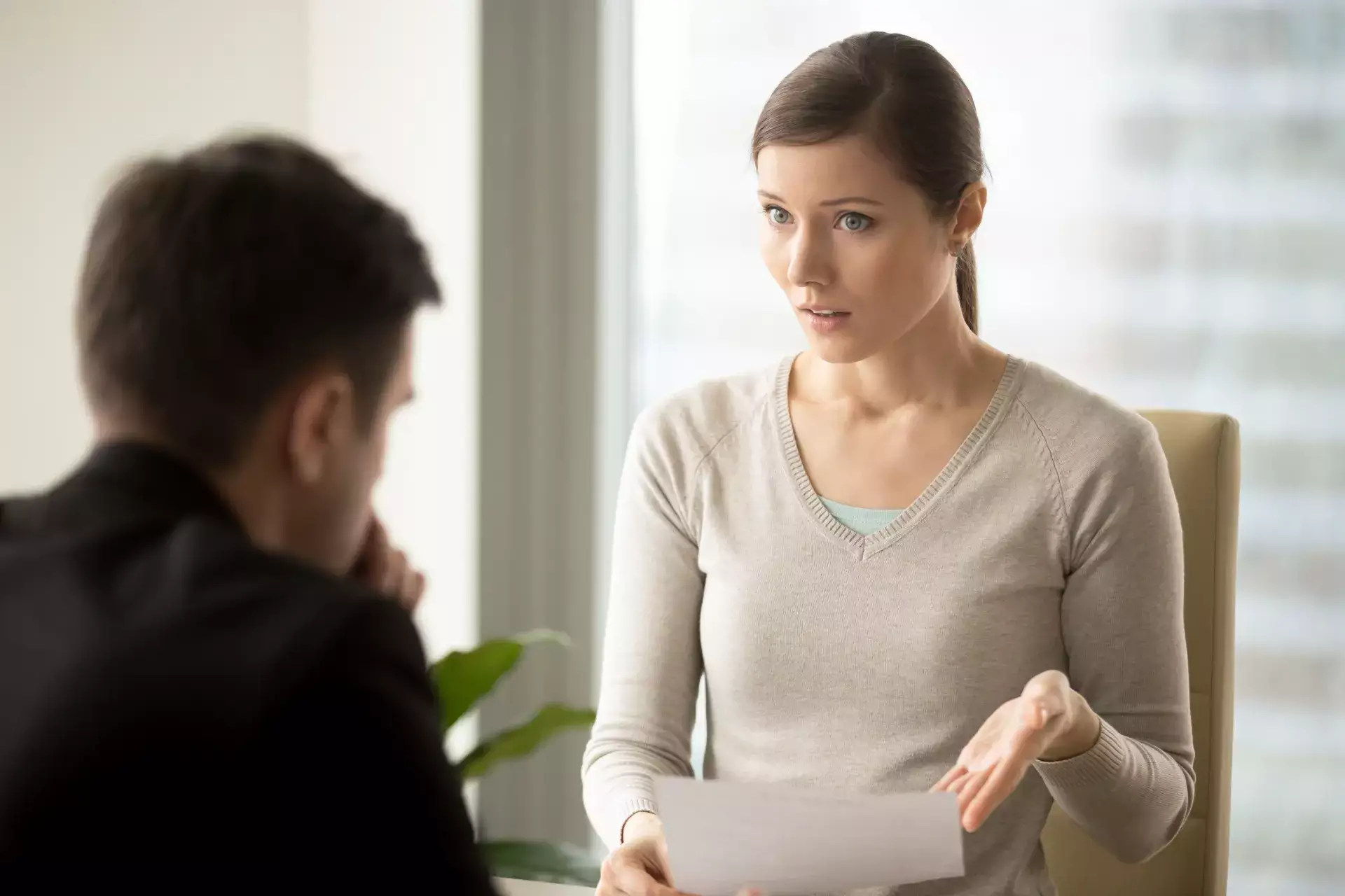 Mulher em ambiente corporativo conversando com um colega, gesticulando durante uma conversa de feedback no trabalho, com ambos sentados em frente a janelas desfocadas.