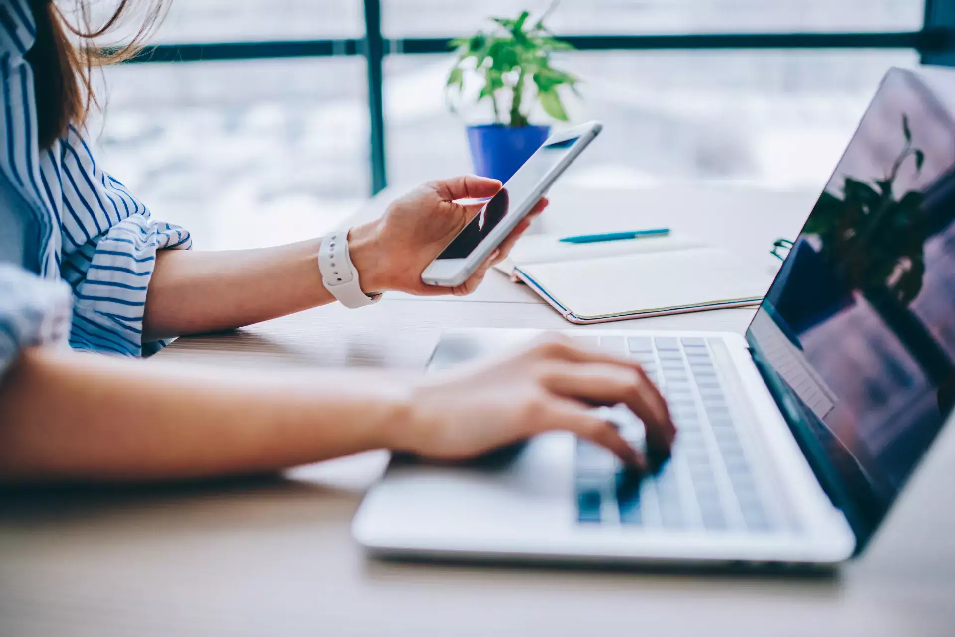 Mulher usando smartphone enquanto trabalha ao lado de um notebook em mesa de escritório, com cadernos e canetas, ambiente corporativo para informática básica para trabalho
